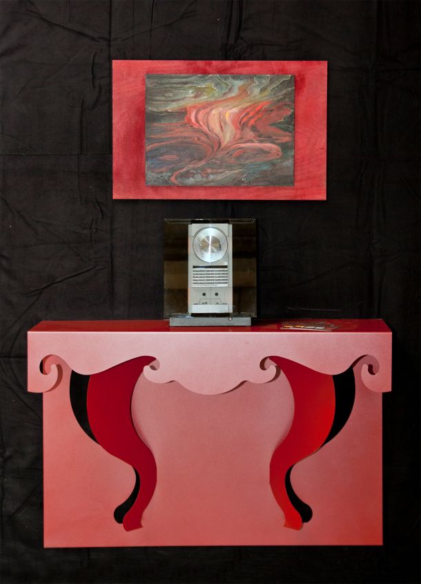 Roter Design-Konsolentisch aus gelasertem Metall mit geschwungenen Formen, Technikobjekt und abstraktem Gemälde auf schwarzem Hintergrund.